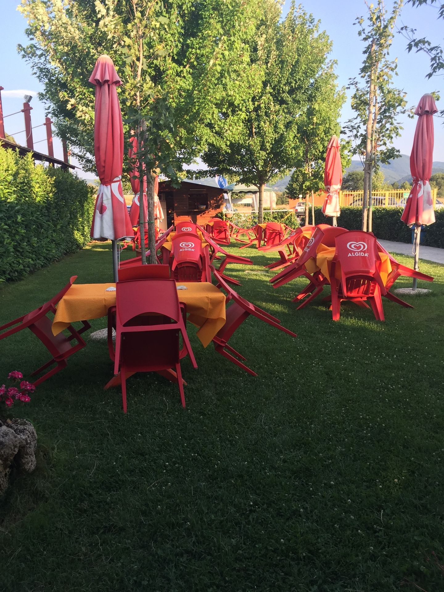 Outdoor cafe area with red chairs and umbrellas overturned on green grass under trees.