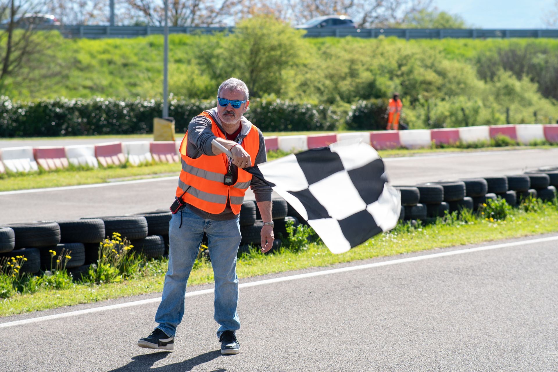 Race official waving checkered flag at a racetrack, wearing a safety vest and sunglasses.