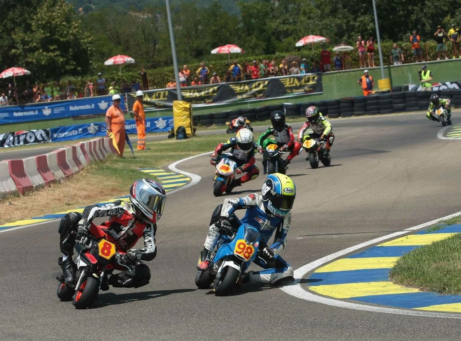 Motorcycle racers on mini bikes competing on a curved track with spectators and officials in the background.