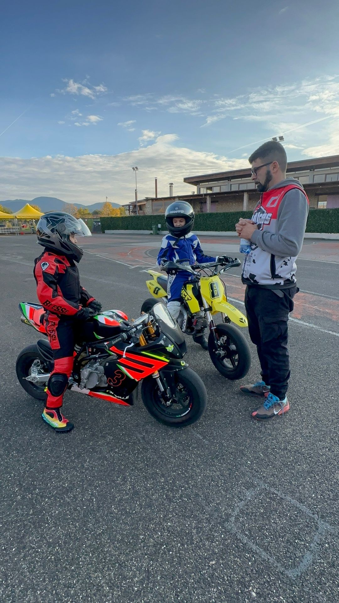 Two children in racing suits and helmets on mini motorcycles talking to an adult at an outdoor track.
