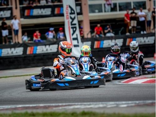 Go-kart drivers racing on a track with spectators in the background during a competitive event.