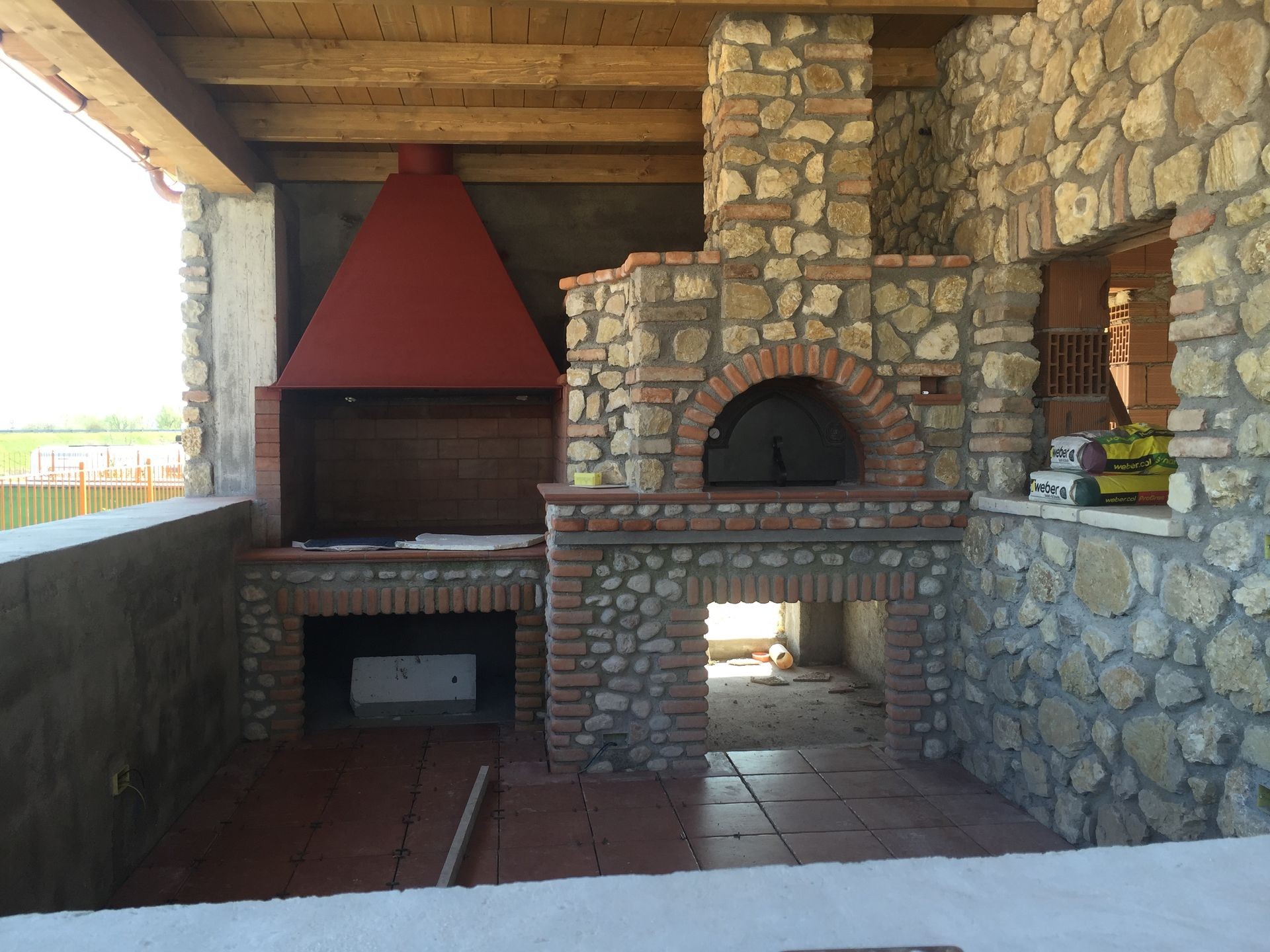 Outdoor stone oven and grill with a red chimney under a wooden roof.