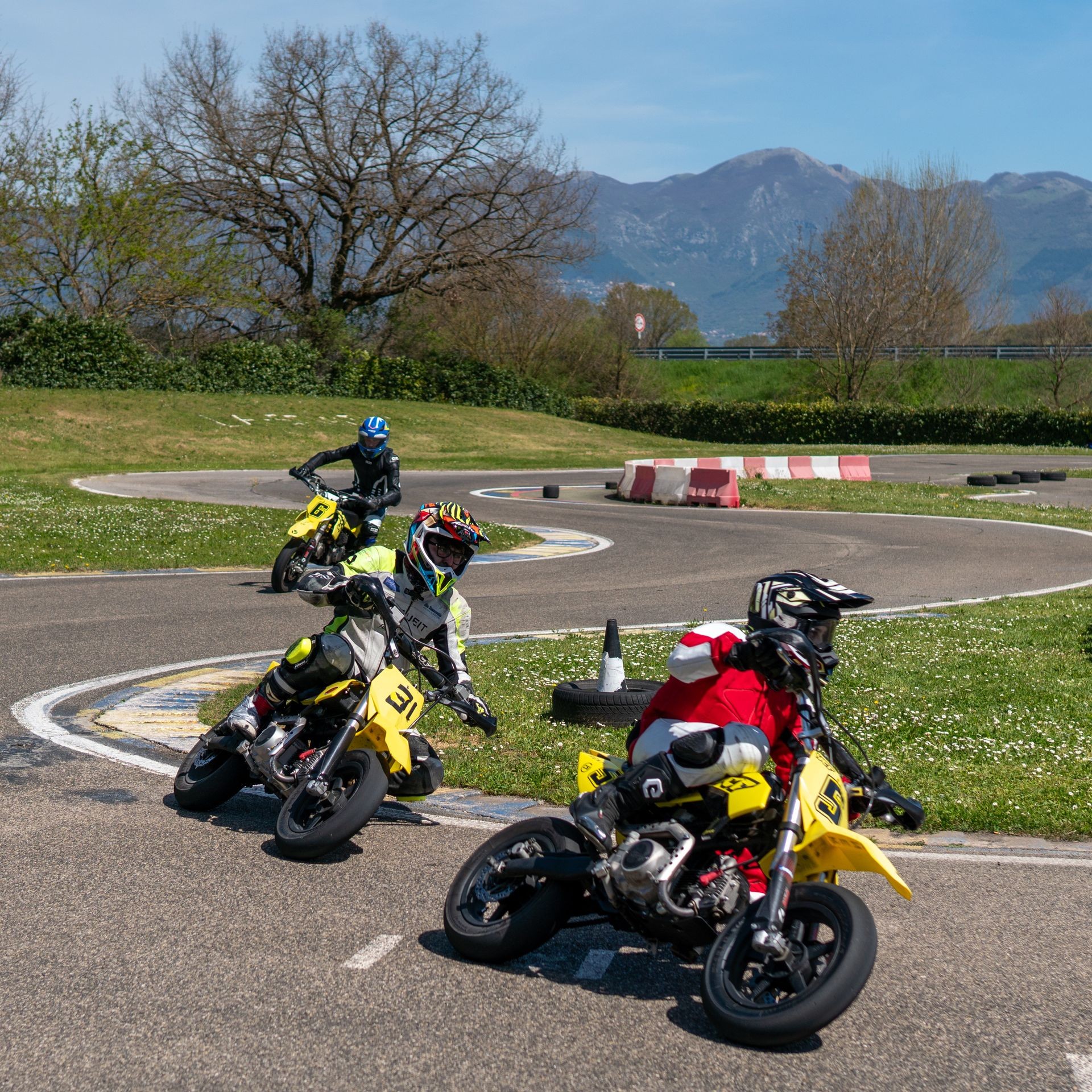 Three motorcyclists in vibrant gear racing on a curvy track with mountains in the background.