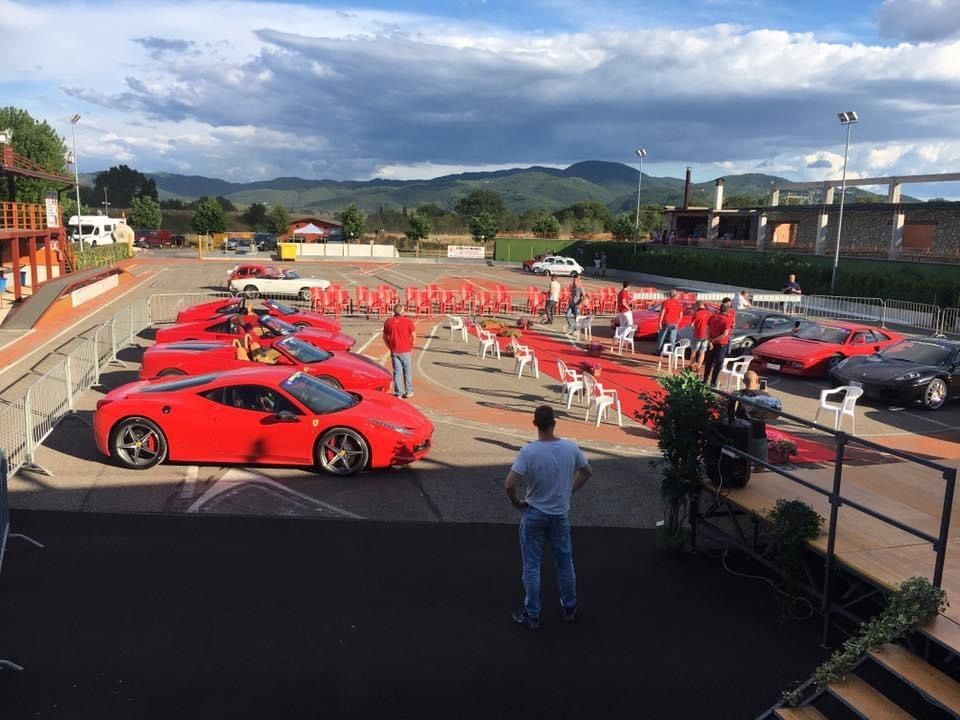 Outdoor car show featuring multiple red sports cars, with people standing around and mountains in the background.